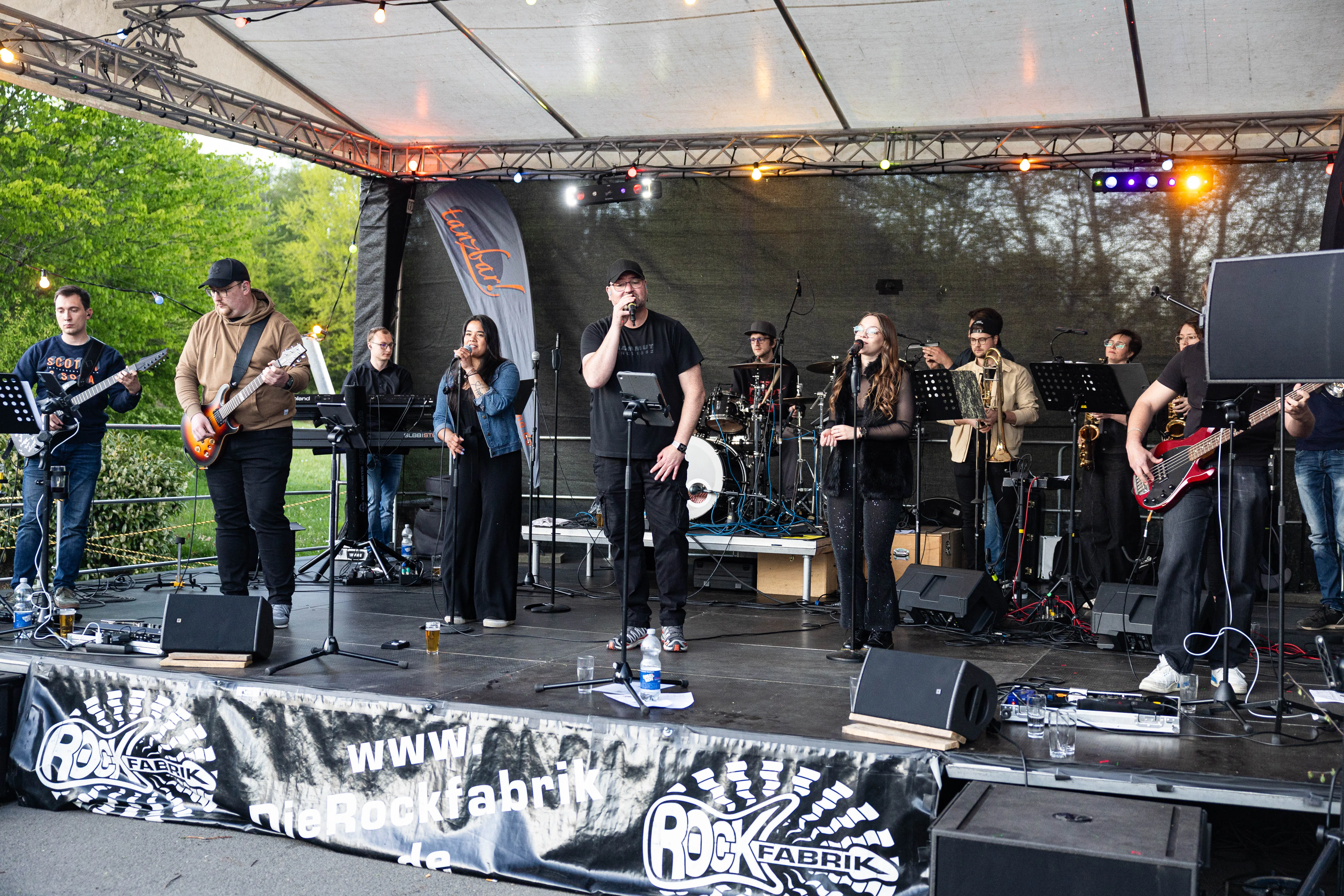 Eine Band spielt auf einer Bühne im Freien, vor ihnen steht ein Sänger mit Kappe und Mikrofon. Im Hintergrund ein Banner mit der Aufschrift „www.dierockfabrik.de“.