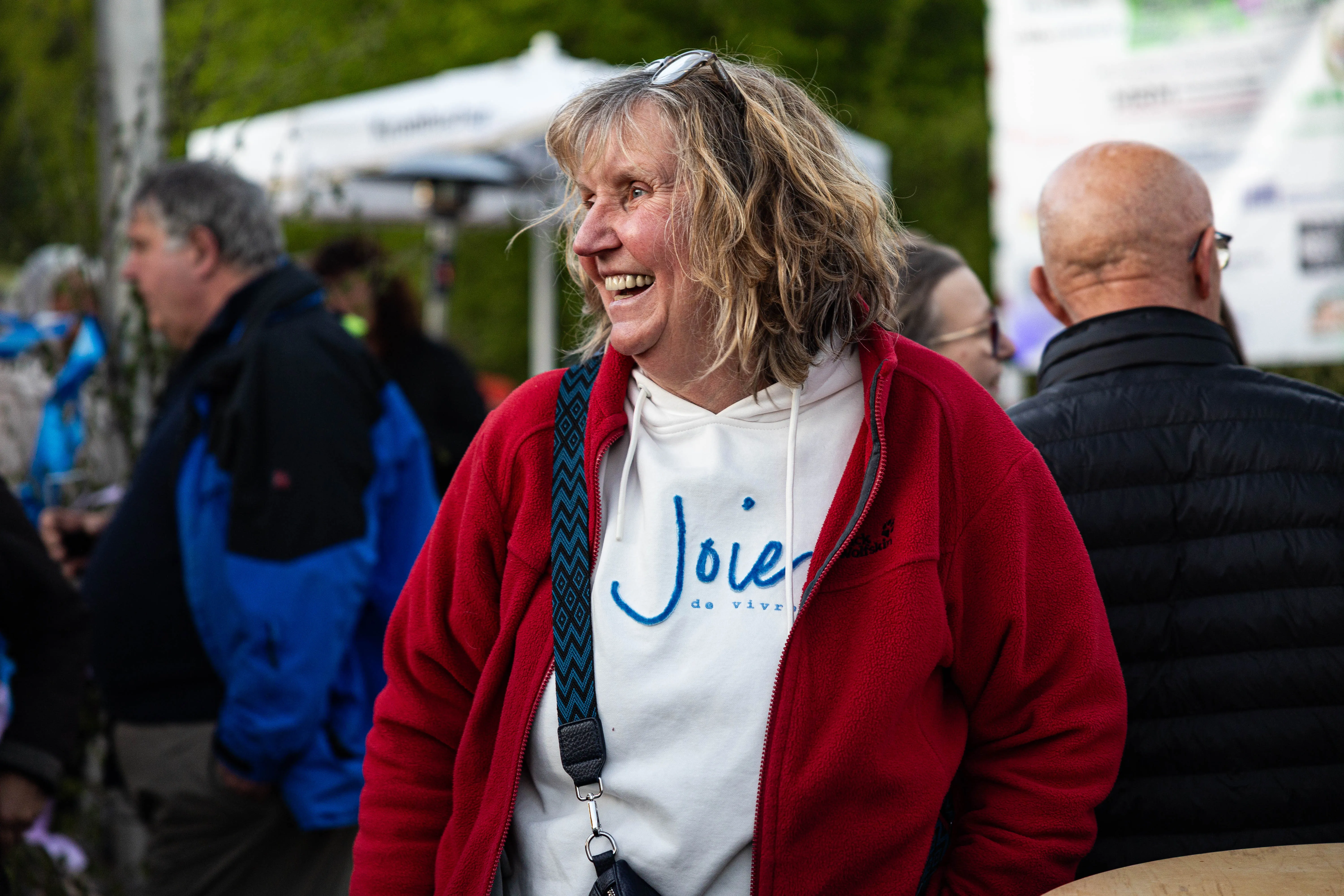 Lachende Frau mit lockigem Haar und rotem Fleecejackett trägt ein weißes Shirt mit dem Aufdruck „Joie de vivre“ auf einem Volksfest.