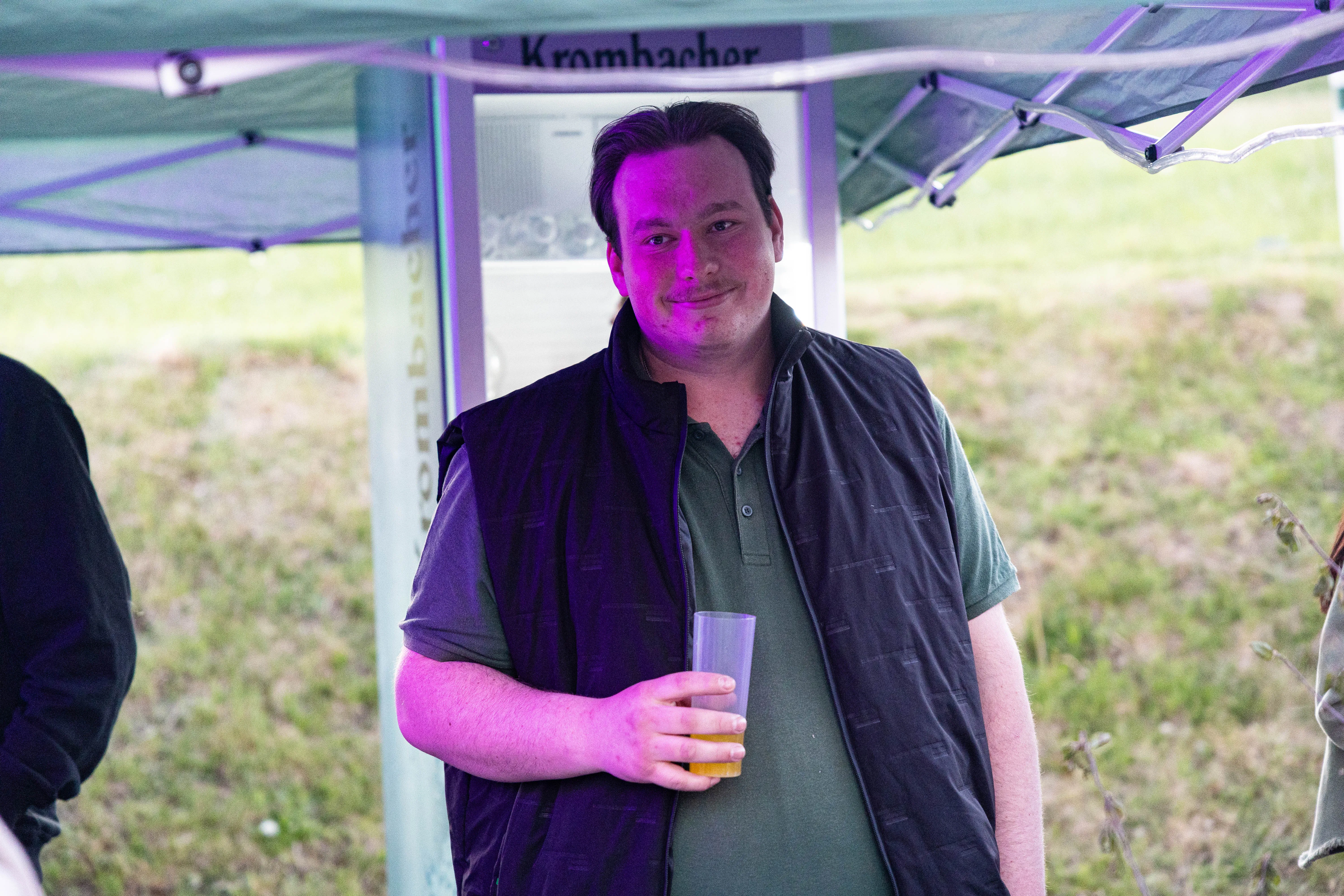 Ein Mann mit dunklem Haar und grünem Poloshirt lächelt in die Kamera, während er ein Glas Bier hält. Im Hintergrund ein Stand mit der Aufschrift „Krombacher“ und ein Zelt.