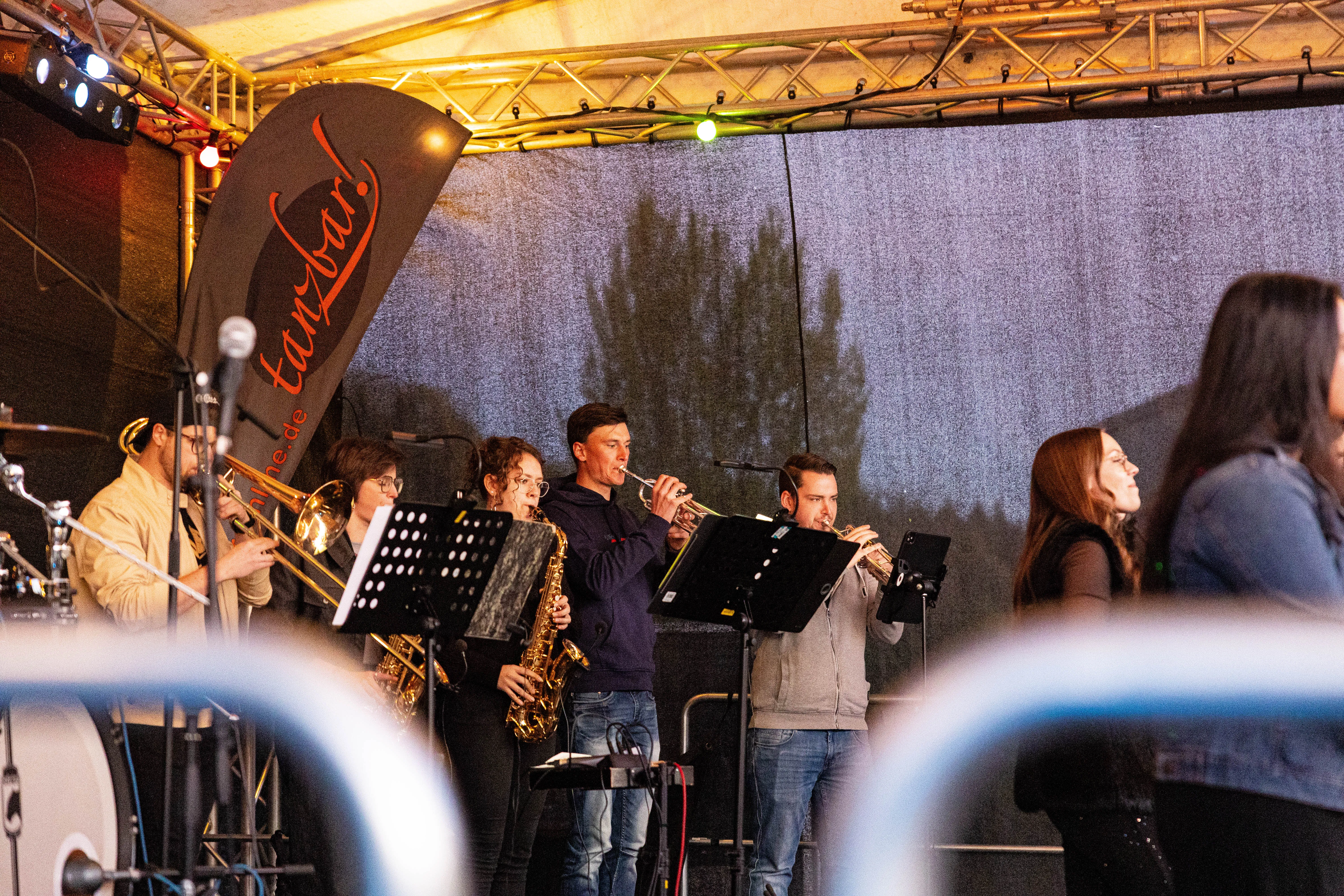 Mehrere Musiker spielen Blasinstrumente auf einer Bühne unter einem Banner mit der Aufschrift „tanzbar!“. Im Hintergrund sind Bäume und ein dunkler Himmel zu sehen.