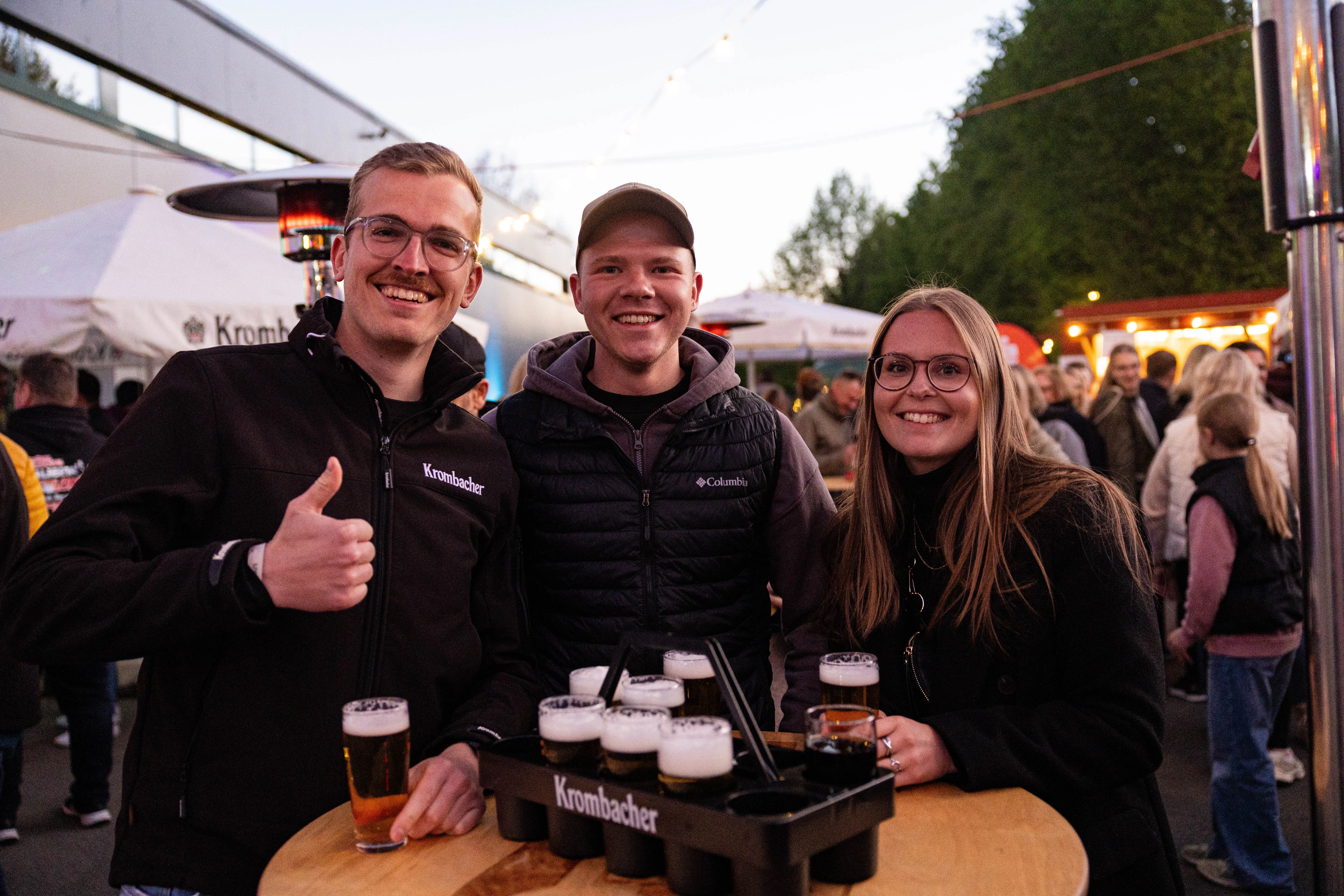 Drei junge Erwachsene lächeln in die Kamera, zwei Männer und eine Frau stehen vor einem Tisch mit Biergläsern. Im Hintergrund sind Menschen auf einem Volksfest zu sehen.
