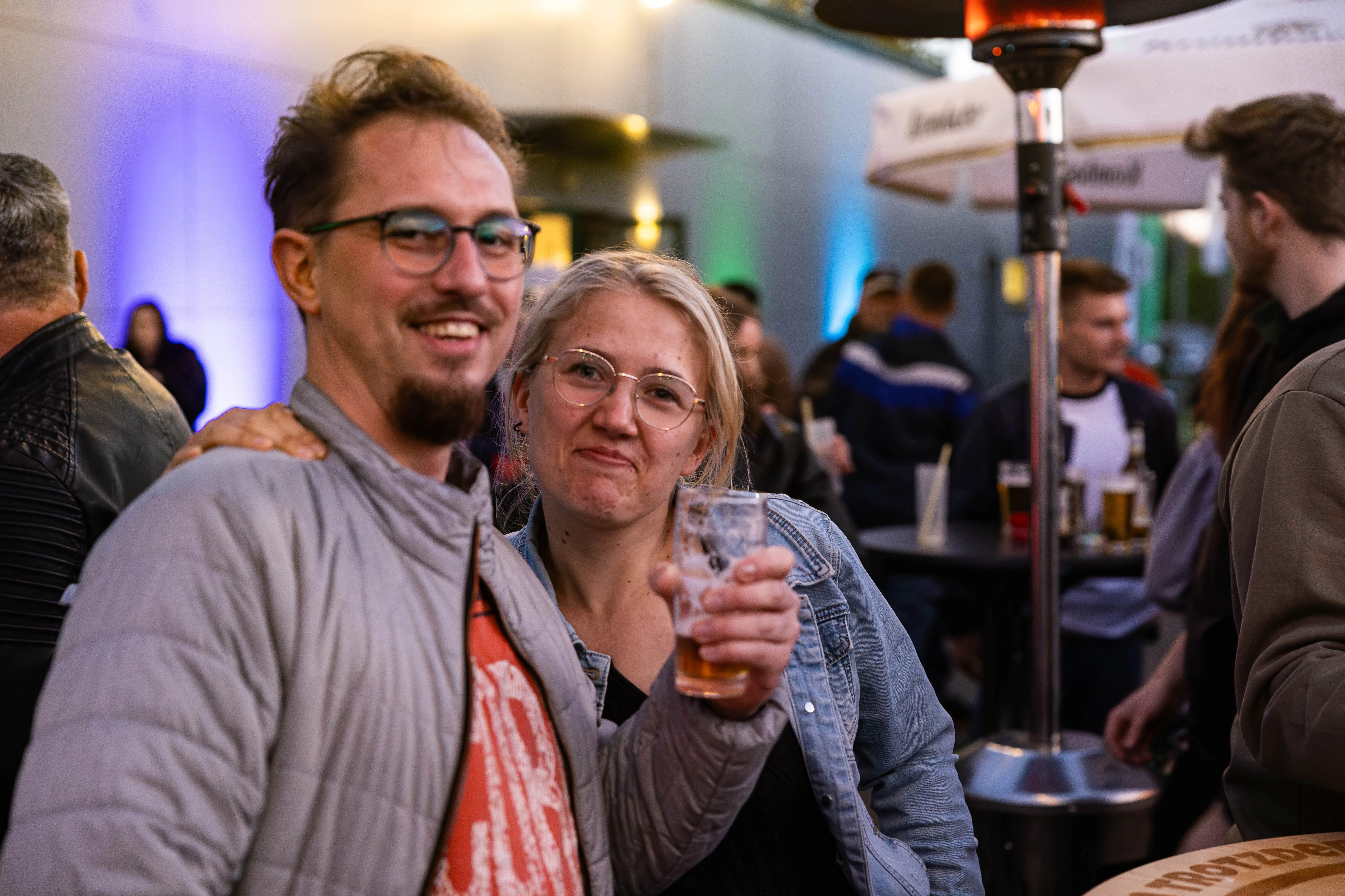 Ein Mann und eine Frau lächeln in die Kamera, die Frau hält ein Bierglas. Im Hintergrund sind weitere Menschen und ein Heizstrahler zu sehen.