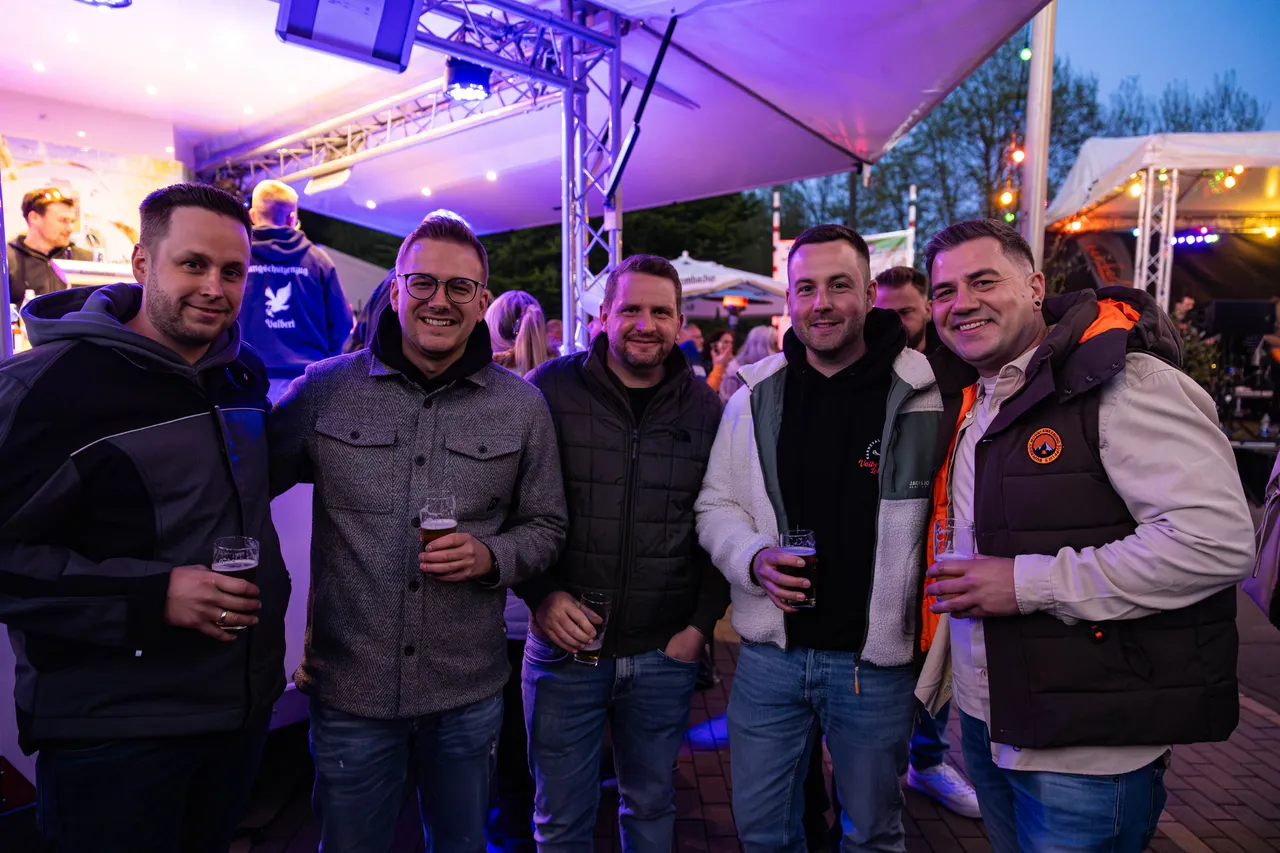 Fünf Männer stehen lächelnd mit Biergläsern vor einer Bühne im Freien, im Hintergrund weitere Menschen und festliche Beleuchtung.