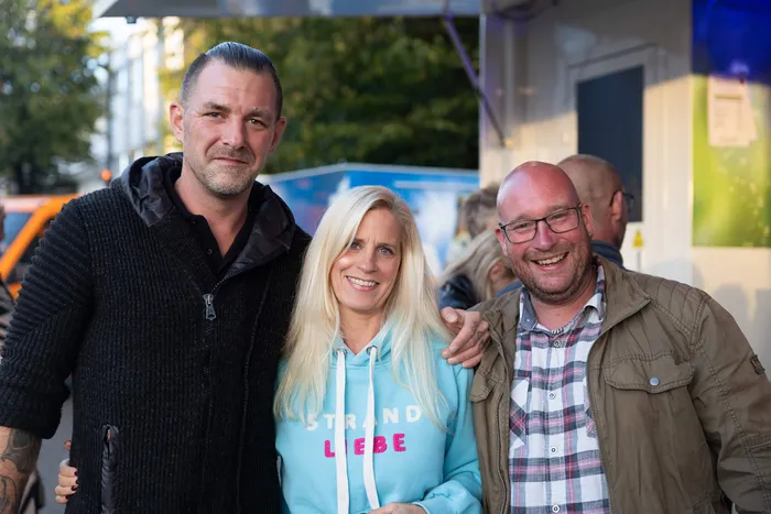Drei lächelnde Personen posieren bei einem Gemeindefest; die Frau in der Mitte trägt einen hellblauen Hoodie mit der Aufschrift „STRAND LIEBE“.