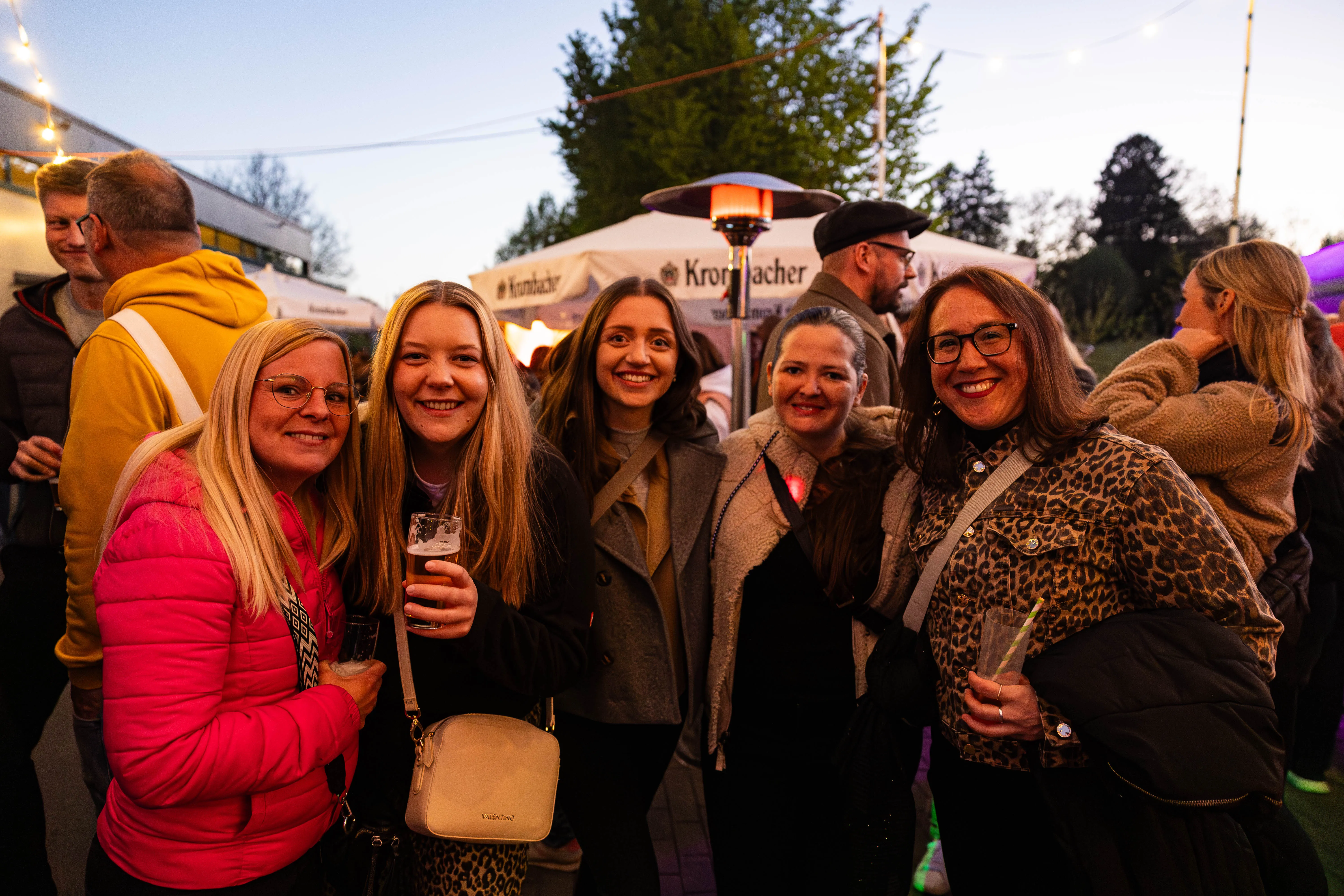 Fünf lächelnde Frauen stehen eng zusammen, einige halten Gläser in der Hand. Im Hintergrund sind weitere Menschen und Zelte zu sehen, vermutlich bei einer Veranstaltung im Freien.