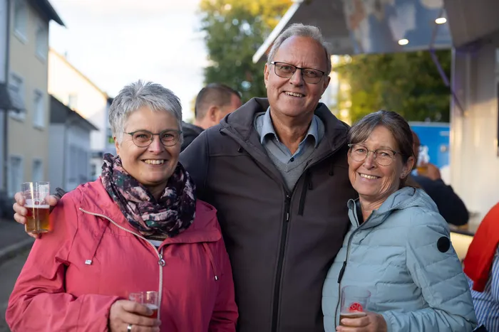Drei lächelnde Personen halten Getränke bei einem Gemeindefest.