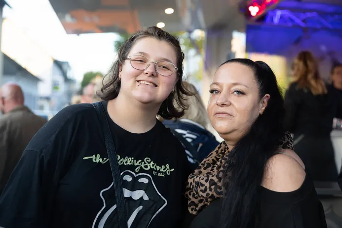 Zwei lächelnde Frauen stehen nebeneinander, eine trägt ein schwarzes Rolling Stones T-Shirt und die andere einen Schal mit Leopardenmuster.