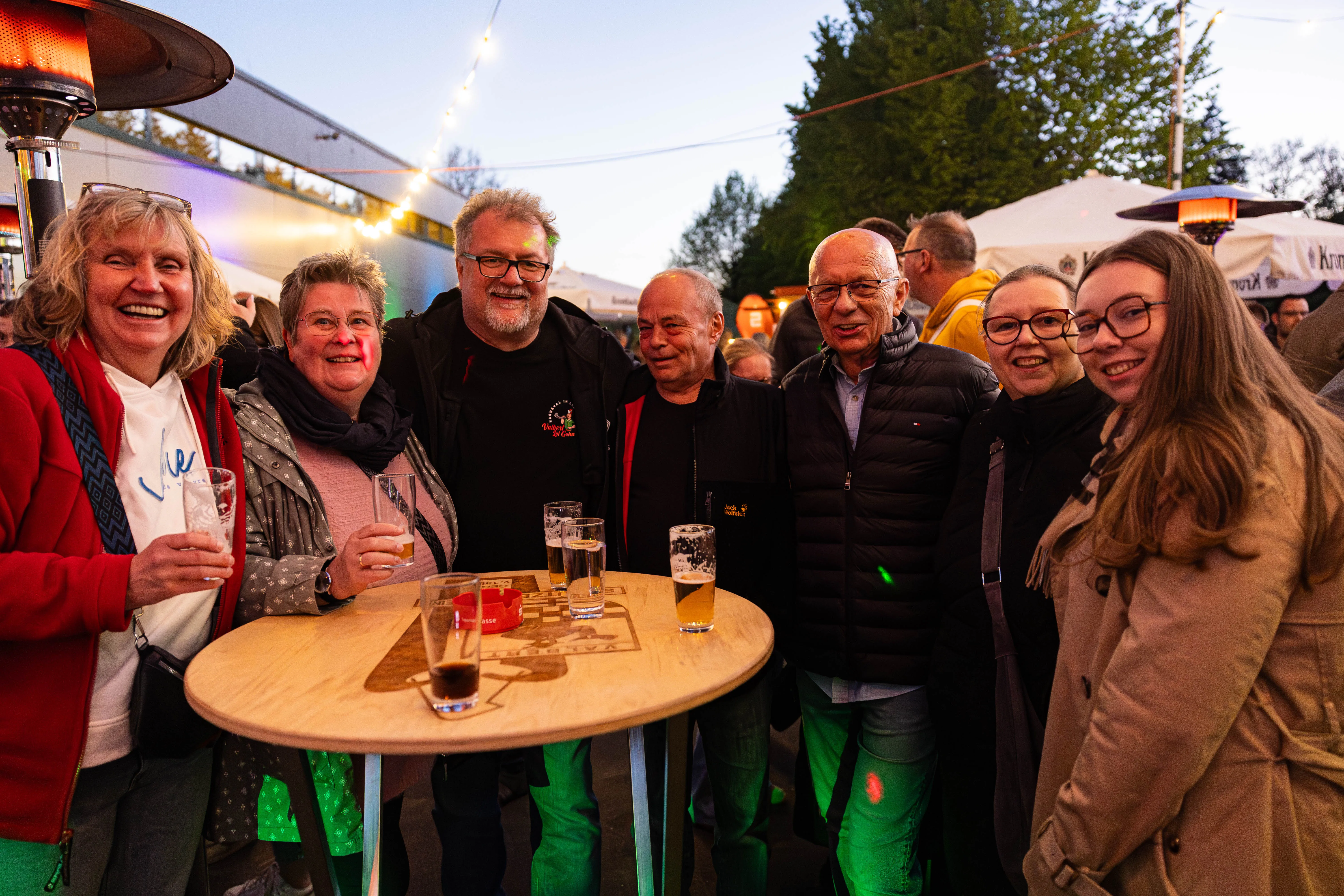 Eine Gruppe von Menschen steht um einen Tisch und hält Gläser mit Getränken in der Hand. Im Hintergrund sind Zelte und Lichter zu sehen.