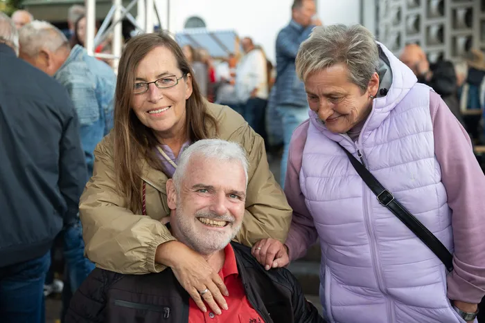 Eine Frau umarmt einen lächelnden Mann, während eine weitere Frau in einer lila Weste daneben steht, bei einem Gemeindefest in Halver.