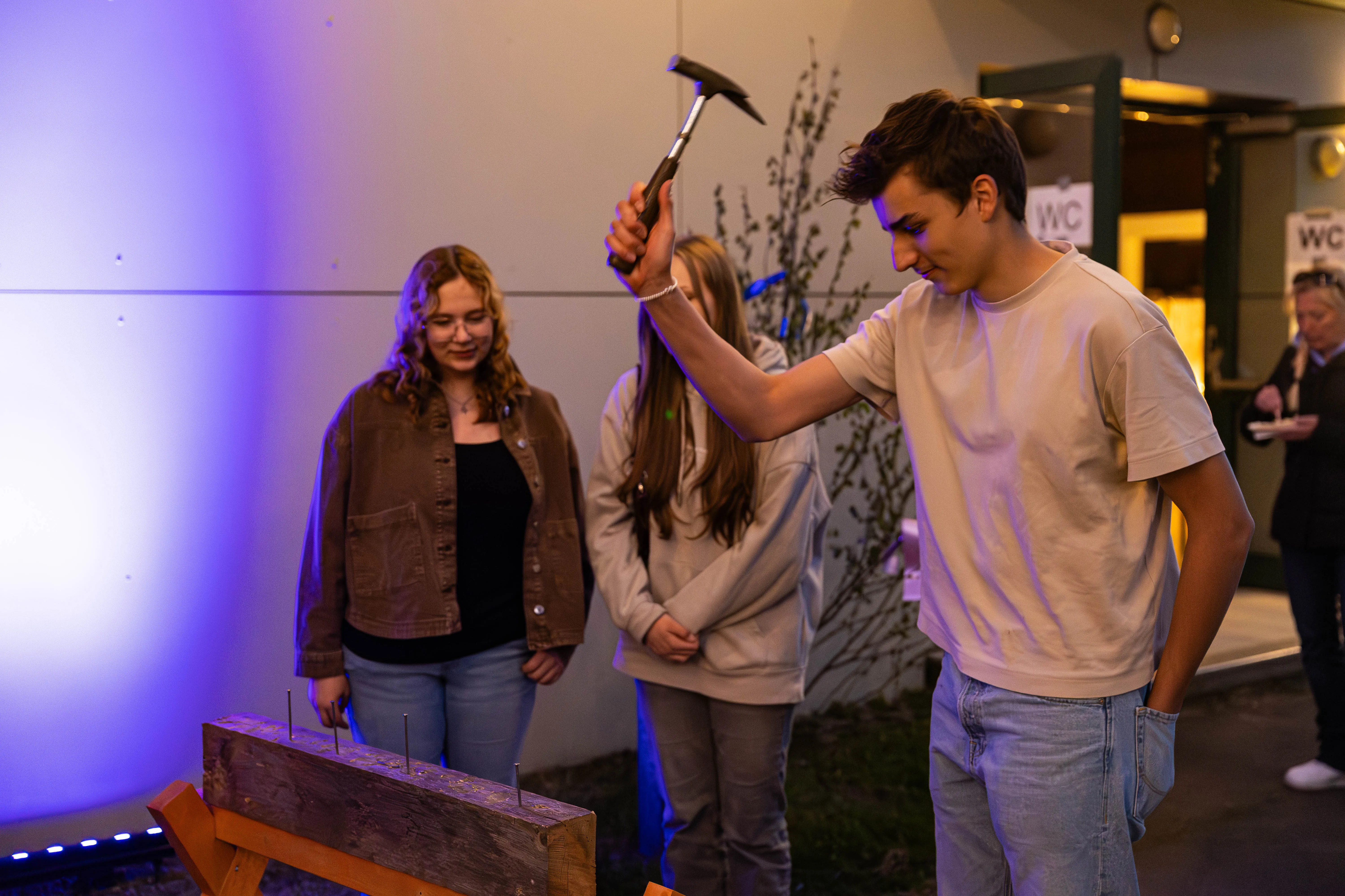 Junger Mann schlägt mit einem Hammer Nägel in ein Holzbrett, während zwei junge Frauen im Hintergrund zusehen. Im Hintergrund sind Schilder mit der Aufschrift „WC“ zu erkennen.