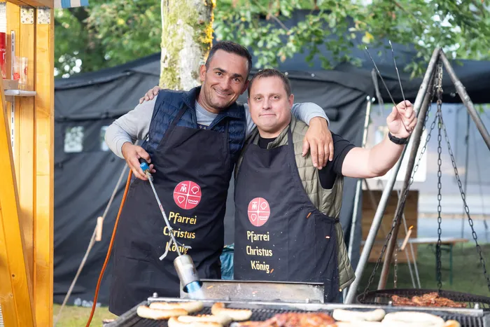 Zwei lächelnde Männer in schwarzen Schürzen der Pfarrei Christus König stehen vor einem Grill mit Fleisch und Würstchen.