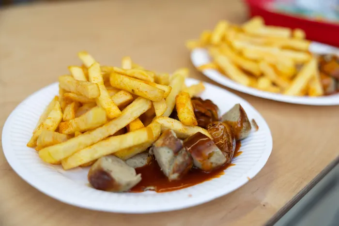 Zwei Pappteller mit Pommes frites und in Sauce geschnittenen Würstchen auf einem Holztisch.