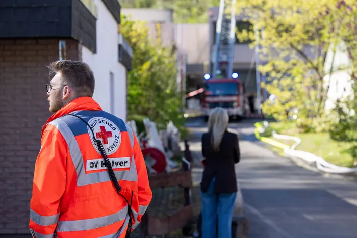 Ein Sanitäter des Deutschen Roten Kreuzes mit Warnweste steht mit dem Rücken zur Kamera, während im Hintergrund Einsatzfahrzeuge und weitere Personen zu sehen sind.