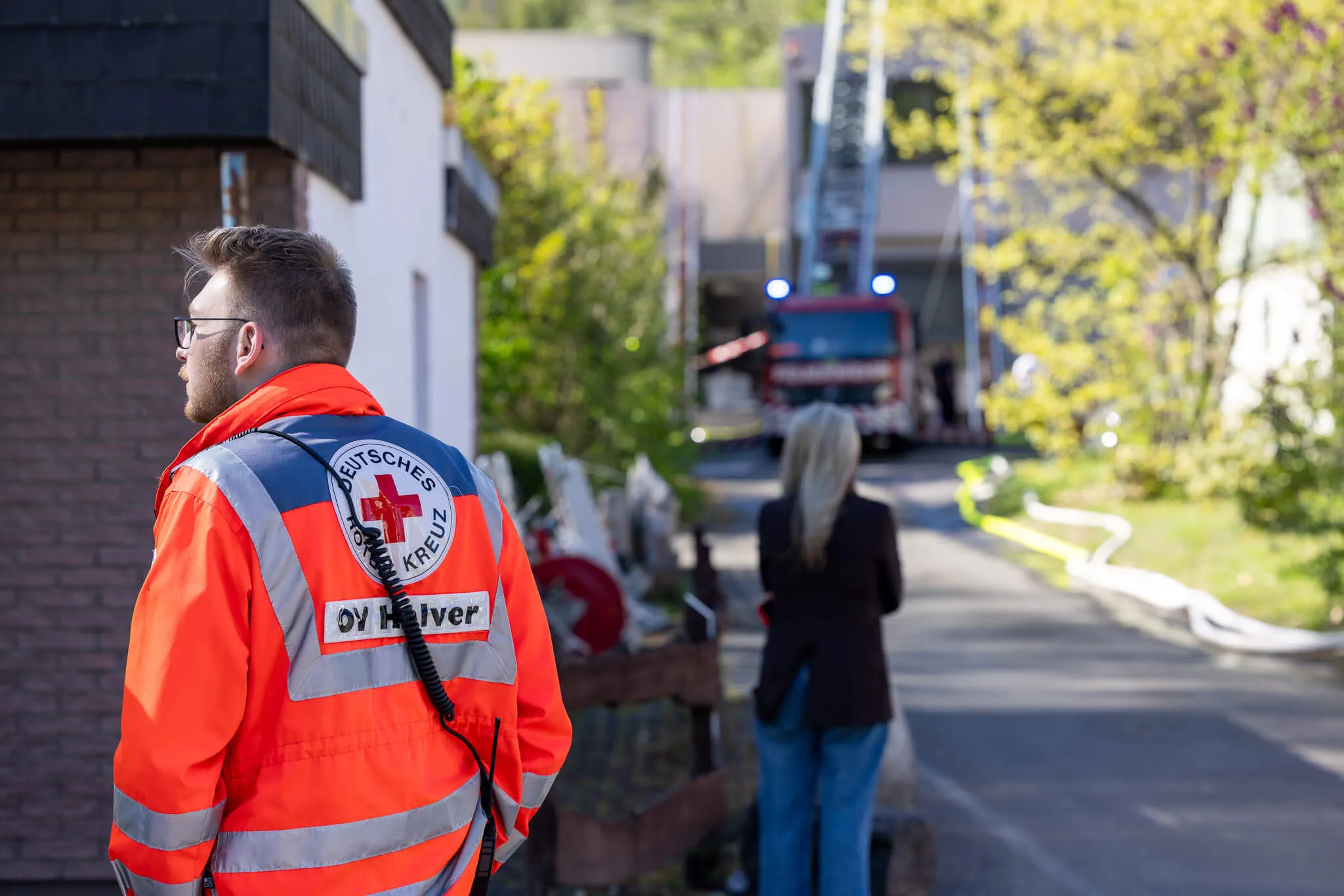 Ein Sanitäter des Deutschen Roten Kreuzes mit Warnweste steht mit dem Rücken zur Kamera, während im Hintergrund Einsatzfahrzeuge und weitere Personen zu sehen sind.