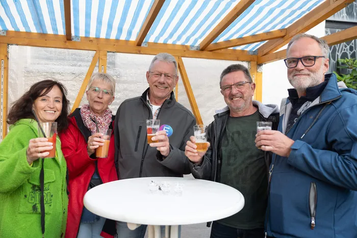 Fünf lächelnde Personen halten Gläser mit Getränken hoch unter einer blau-weiß gestreiften Markise bei einem Gemeindefest.