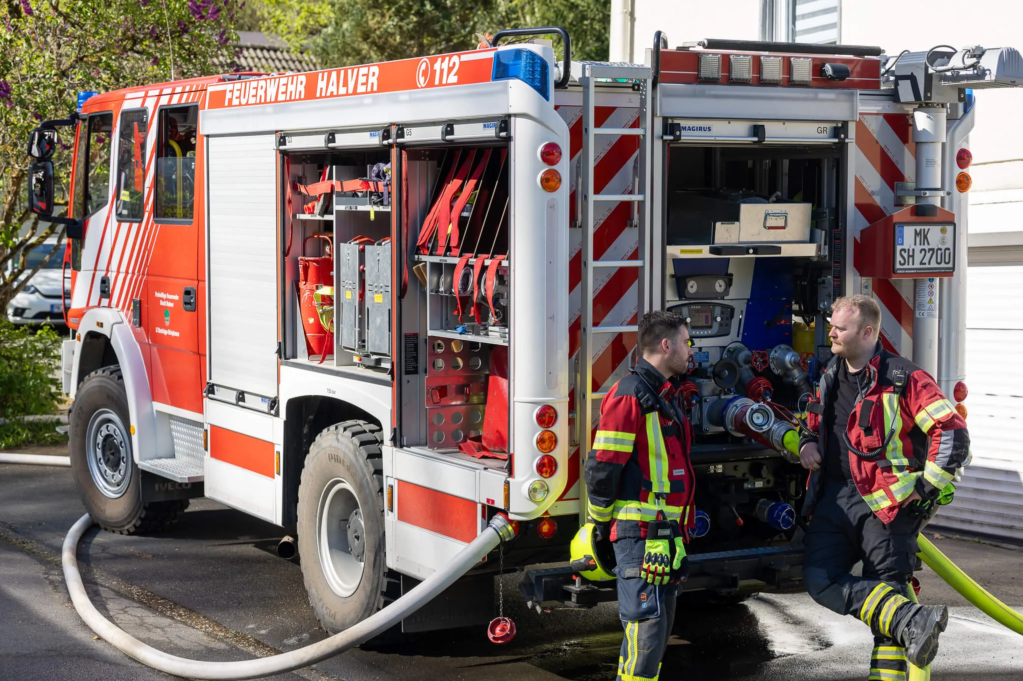 Rotes Feuerwehrauto mit geöffneten Fächern, zwei Feuerwehrleute in voller Montur stehen davor und halten einen Schlauch.
