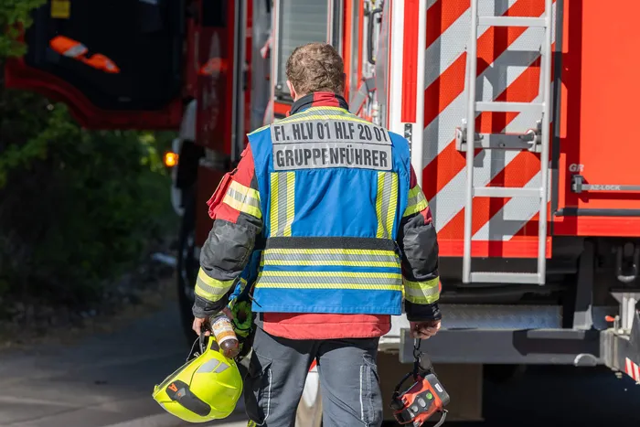 Feuerwehrmann mit der Aufschrift „Gruppenführer“ geht mit zwei Helmen an einem roten Feuerwehrauto vorbei.