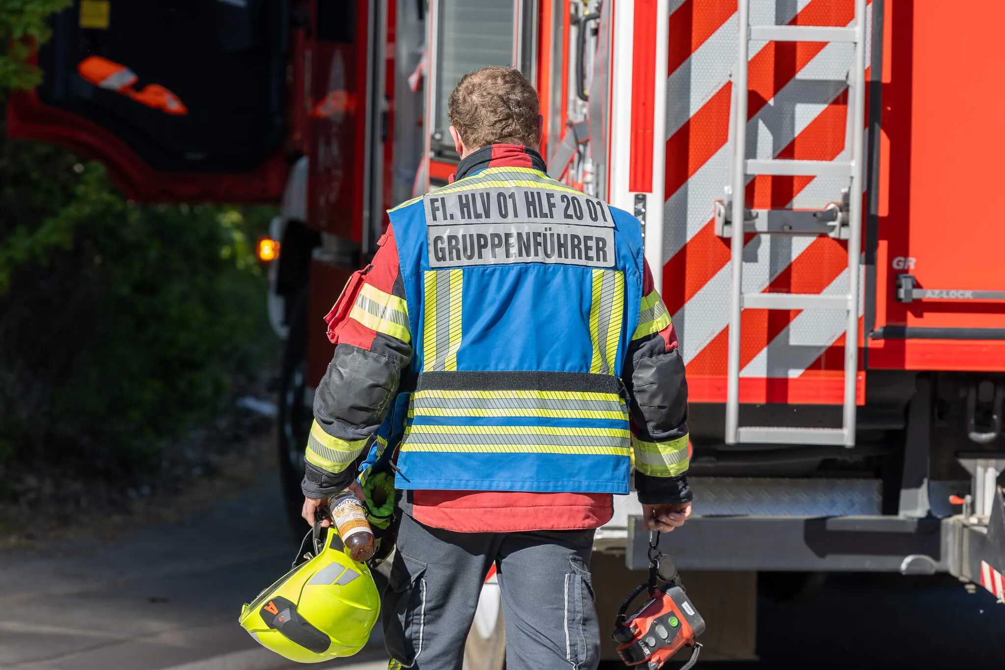 Feuerwehrmann mit der Aufschrift „Gruppenführer“ geht mit zwei Helmen an einem roten Feuerwehrauto vorbei.