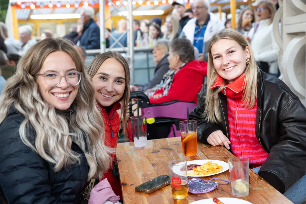Drei lächelnde junge Frauen sitzen an einem Tisch mit Getränken und Speisen bei einem Gemeindefest.