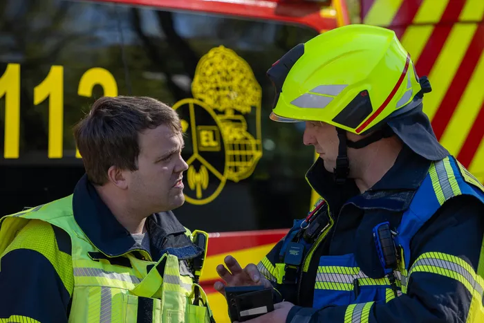 Zwei Männer, einer mit und einer ohne Feuerwehrhelm, stehen sich im Gespräch gegenüber. Im Hintergrund ist die Zahl 112 und ein Feuerwehrabzeichen zu sehen.