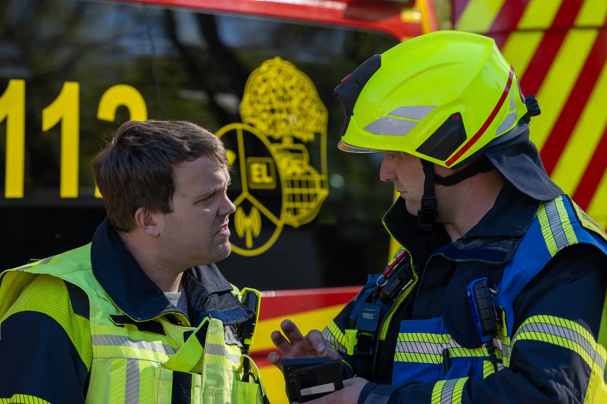 Zwei Männer, einer mit und einer ohne Feuerwehrhelm, stehen sich im Gespräch gegenüber. Im Hintergrund ist die Zahl 112 und ein Feuerwehrabzeichen zu sehen.