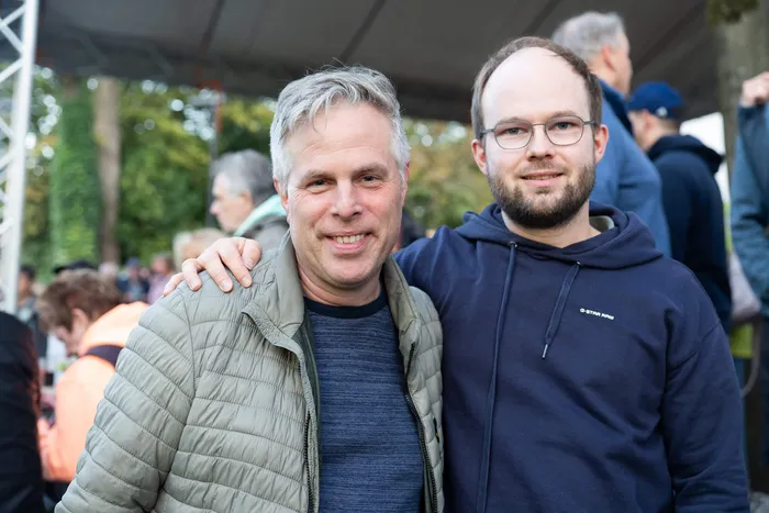 Zwei lächelnde Männer posieren bei einem Gemeindefest; einer trägt eine hellgrüne Steppjacke, der andere einen dunkelblauen Kapuzenpullover.