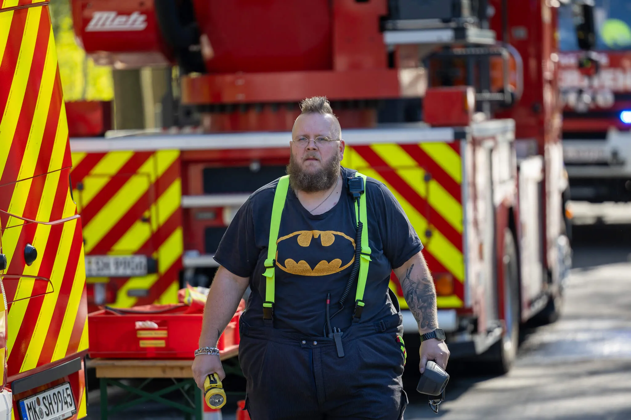 Ein Feuerwehrmann mit Bart und Batman-T-Shirt steht vor einem roten Feuerwehrauto. Im Hintergrund sind weitere Einsatzkräfte und Einsatzfahrzeuge zu sehen.