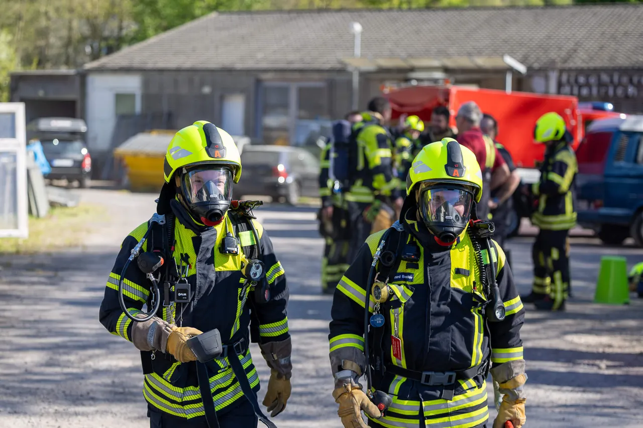 Zwei Feuerwehrleute mit Atemschutzmasken und Einsatzkleidung stehen im Vordergrund, während weitere Einsatzkräfte im Hintergrund arbeiten. Im Hintergrund sind Fahrzeuge und Gebäude zu sehen.
