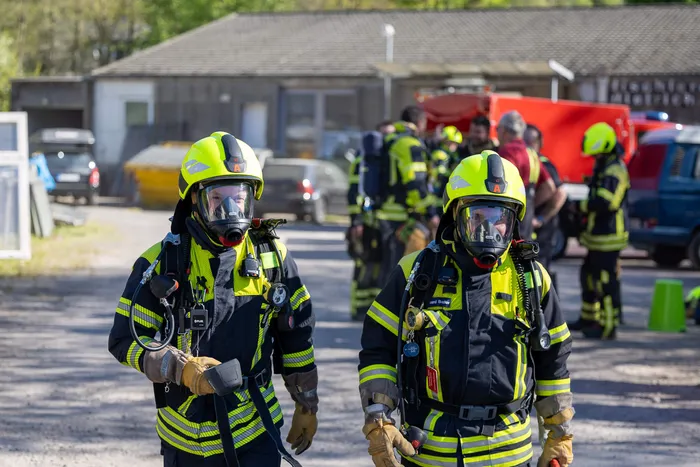 Zwei Feuerwehrleute mit Atemschutzmasken und Einsatzkleidung stehen im Vordergrund, während weitere Einsatzkräfte im Hintergrund arbeiten. Im Hintergrund sind Fahrzeuge und Gebäude zu sehen.