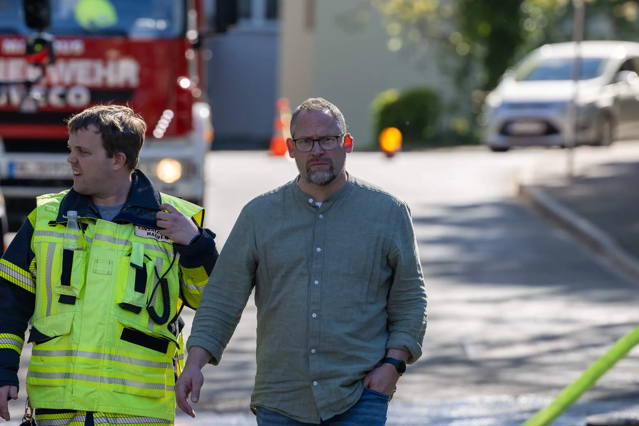 Ein Feuerwehrmann in Warnweste und ein Mann mit Brille gehen neben einem roten Feuerwehrauto auf einer Straße.