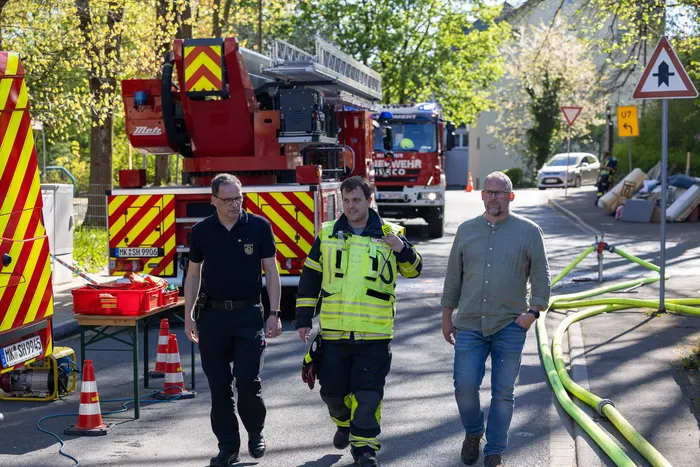 Drei Männer in Anzügen gehen an einem Feuerwehrauto und ausgelegten Löschschläuchen vorbei. Im Hintergrund sind weitere Einsatzkräfte und Fahrzeuge zu sehen.