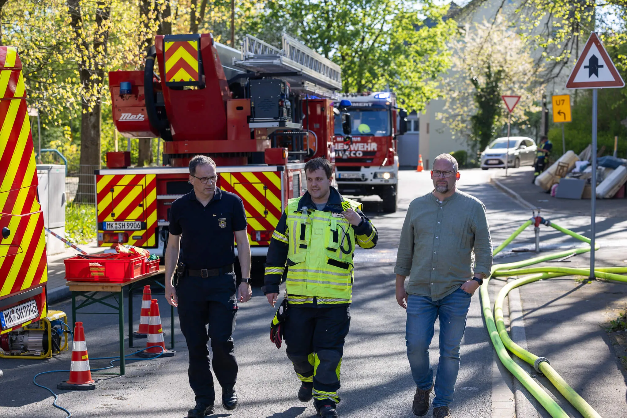Drei Männer in Anzügen gehen an einem Feuerwehrauto und ausgelegten Löschschläuchen vorbei. Im Hintergrund sind weitere Einsatzkräfte und Fahrzeuge zu sehen.