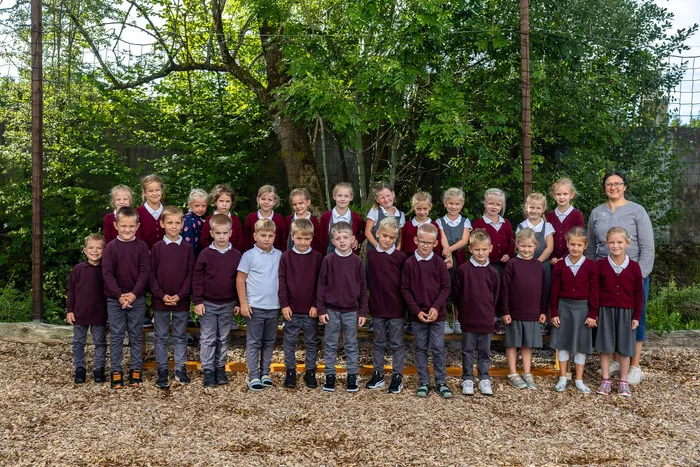 Eine Gruppe von Kindern in weinroten und grauen Schuluniformen posiert gemeinsam mit einer erwachsenen Frau vor Bäumen.