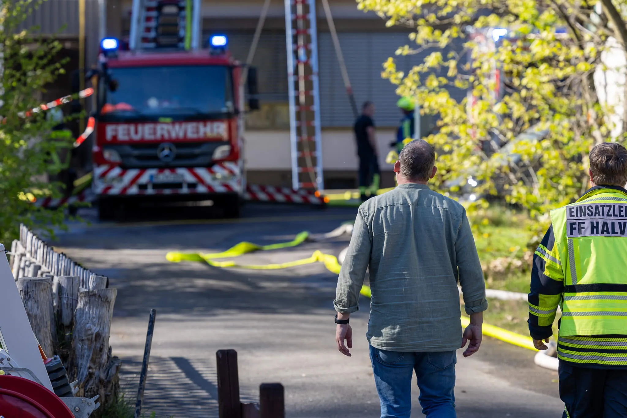 Ein Feuerwehrauto steht vor einem Gebäude, während im Vordergrund zwei Männer mit Warnwesten den Einsatzort betrachten.
