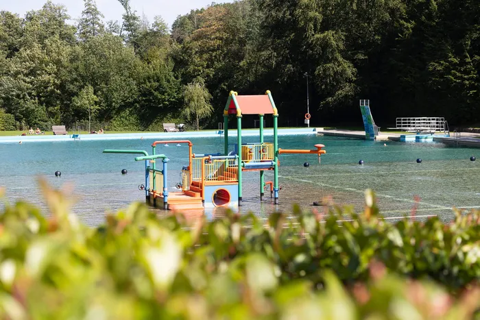 Buntes Wasserspielgerät in einem Freibad vor einer Kulisse aus dichten Bäumen.