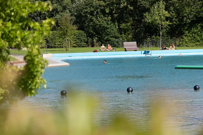 Menschen schwimmen und sonnen sich am Rand eines blauen Außenpools im Waldfreibad Herpine vor einer Waldkulisse.