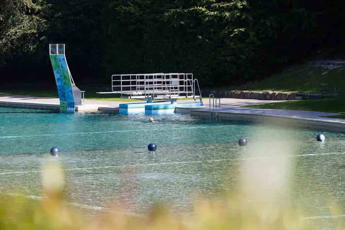 Außenpool mit blauer Wasserrutsche, Sprungturm und blauen Bojen vor einem Waldhintergrund.