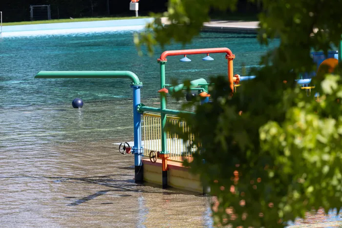 Bunte Wasserspielgeräte mit grünen Rohren und einer orangen Duschanlage in einem flachen Schwimmbecken.