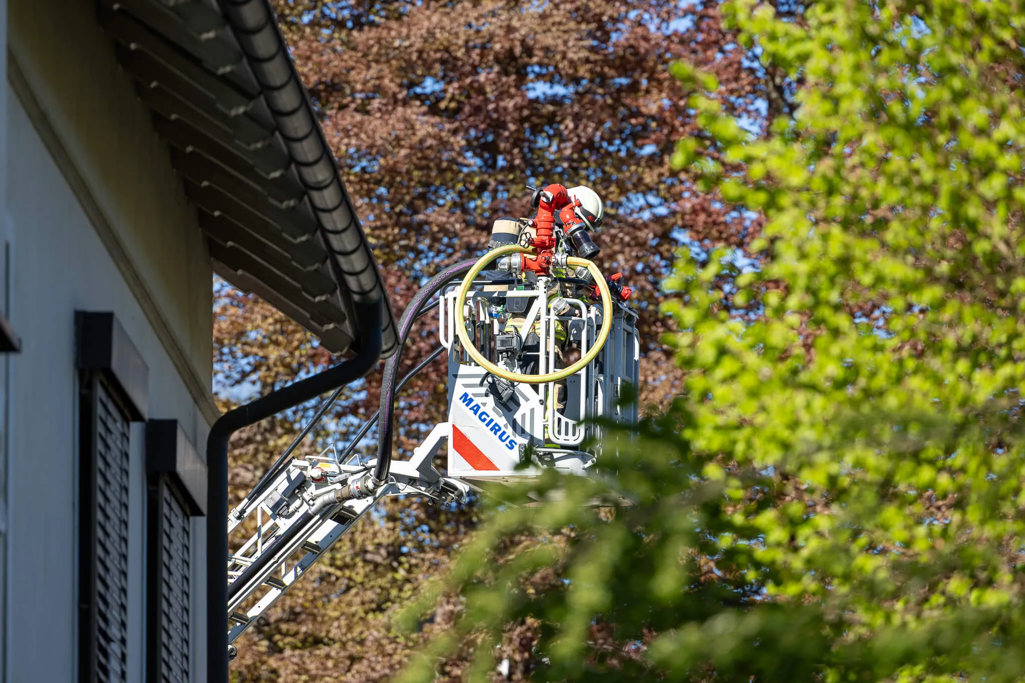 Feuerwehrmann in einer Hebebühne arbeitet an einem Dach, während im Hintergrund ein Baum mit frischem Laub zu sehen ist.