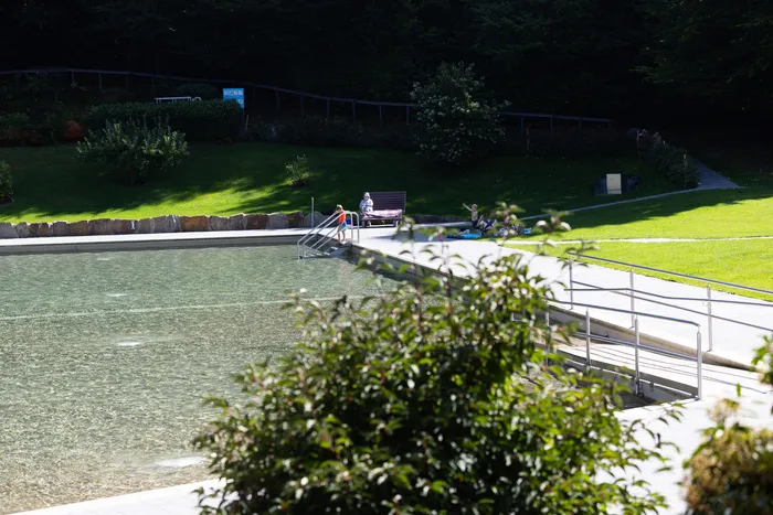 Blick auf das Wasser des Waldfreibads Herpine mit einer Metallreling und Personen auf der angrenzenden Rasenfläche.