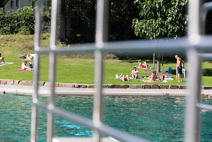 Blick durch einen weißen Metallzaun auf ein Schwimmbecken und Personen, die auf einer Rasenfläche daneben sonnen.