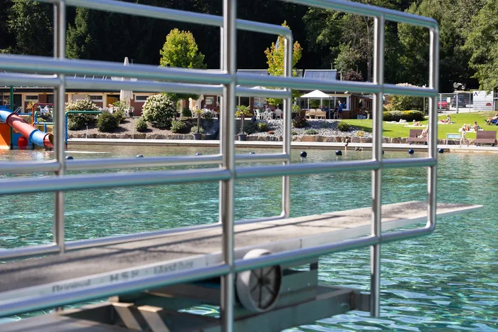 Blick durch ein Metallgeländer auf das türkisfarbene Wasser und einen Steg im Waldfreibad Herpine mit Gebäuden und Bäumen im Hintergrund.