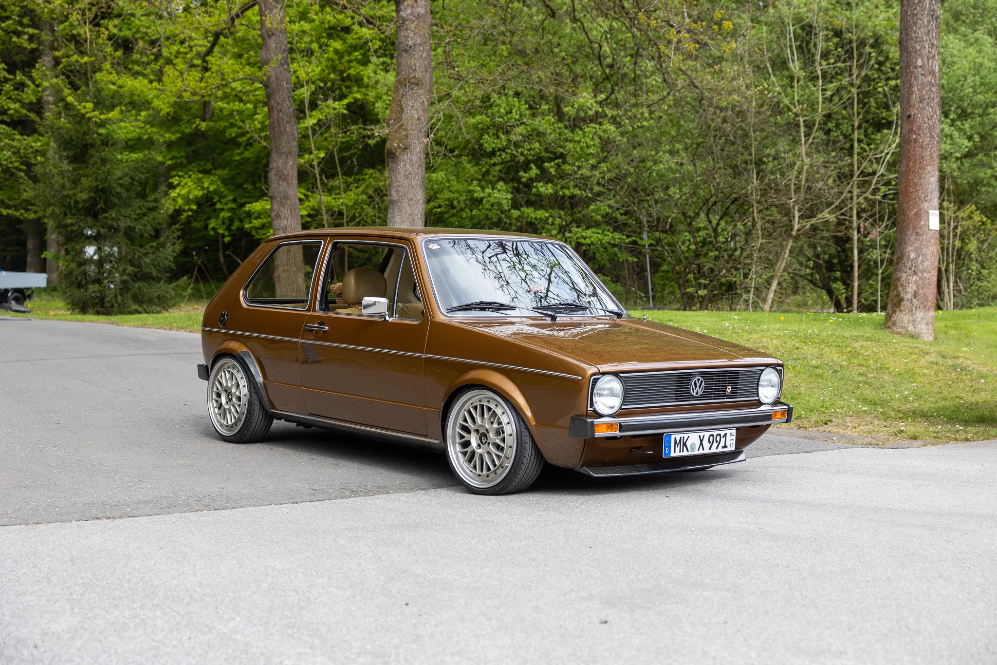 Brauner VW Golf I steht auf einer Straße vor Bäumen, ein Kennzeichen ist sichtbar.