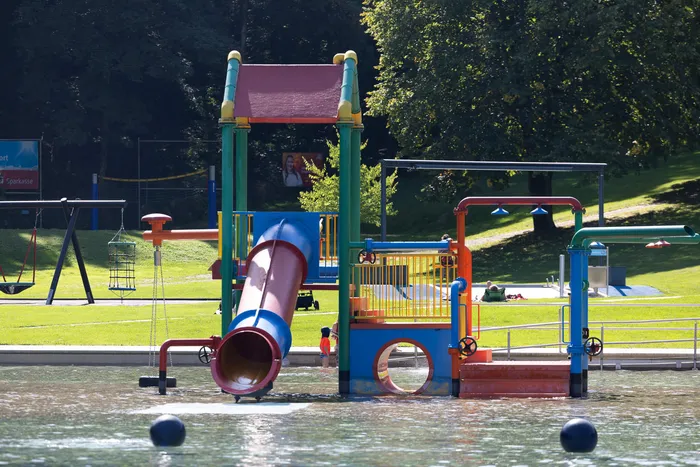 Bunter Wasserspielplatz mit einer roten Röhrenrutsche am Ufer des Waldfreibads Herpine.