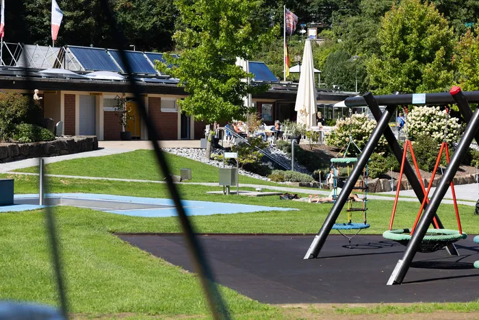 Spielplatz mit Schaukeln und Rasenfläche vor einem Gebäude mit Solaranlagen im Waldfreibad Herpine.