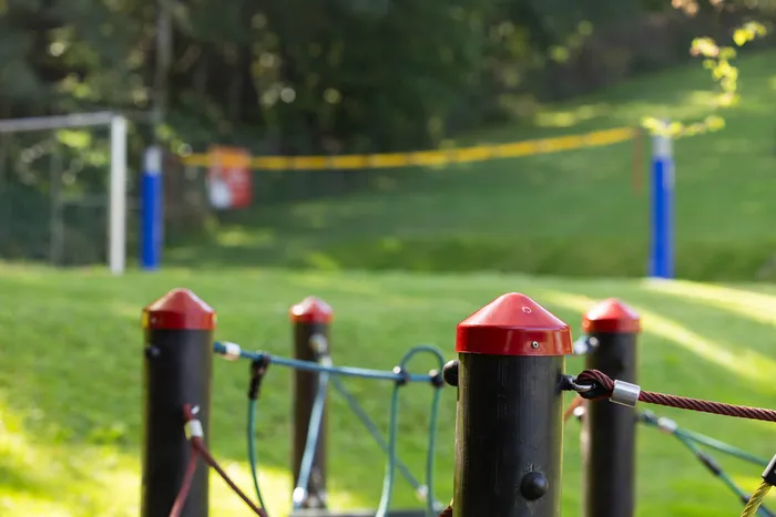 Schwarze Pfosten mit roten Kappen und bunten Seilen vor einer unscharfen Grünanlage im Waldfreibad Herpine.