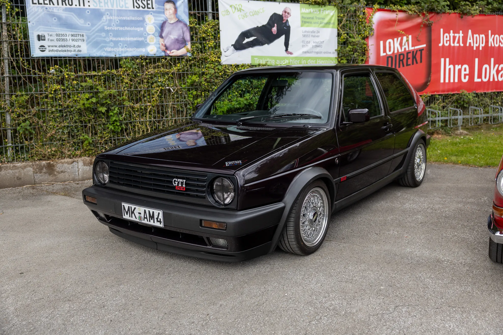 Schwarzer VW Golf GTI der ersten Generation steht auf einem Parkplatz vor einer Hecke. Im Hintergrund ein Banner mit Werbung und ein Mann mit einem Hund.