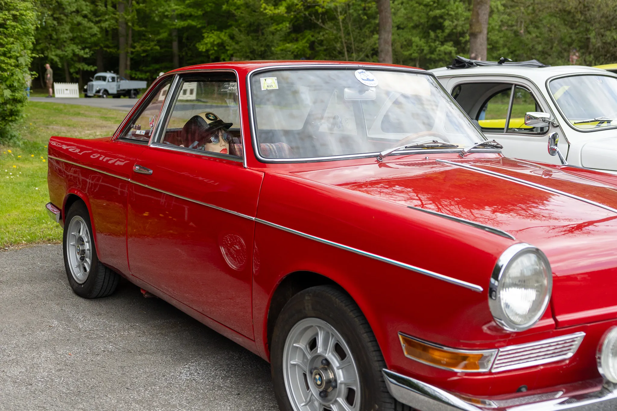 Roter Oldtimer vom Typ Volvo P1800 steht auf einem Parkplatz, ein Mann mit Kappe sitzt am Steuer.