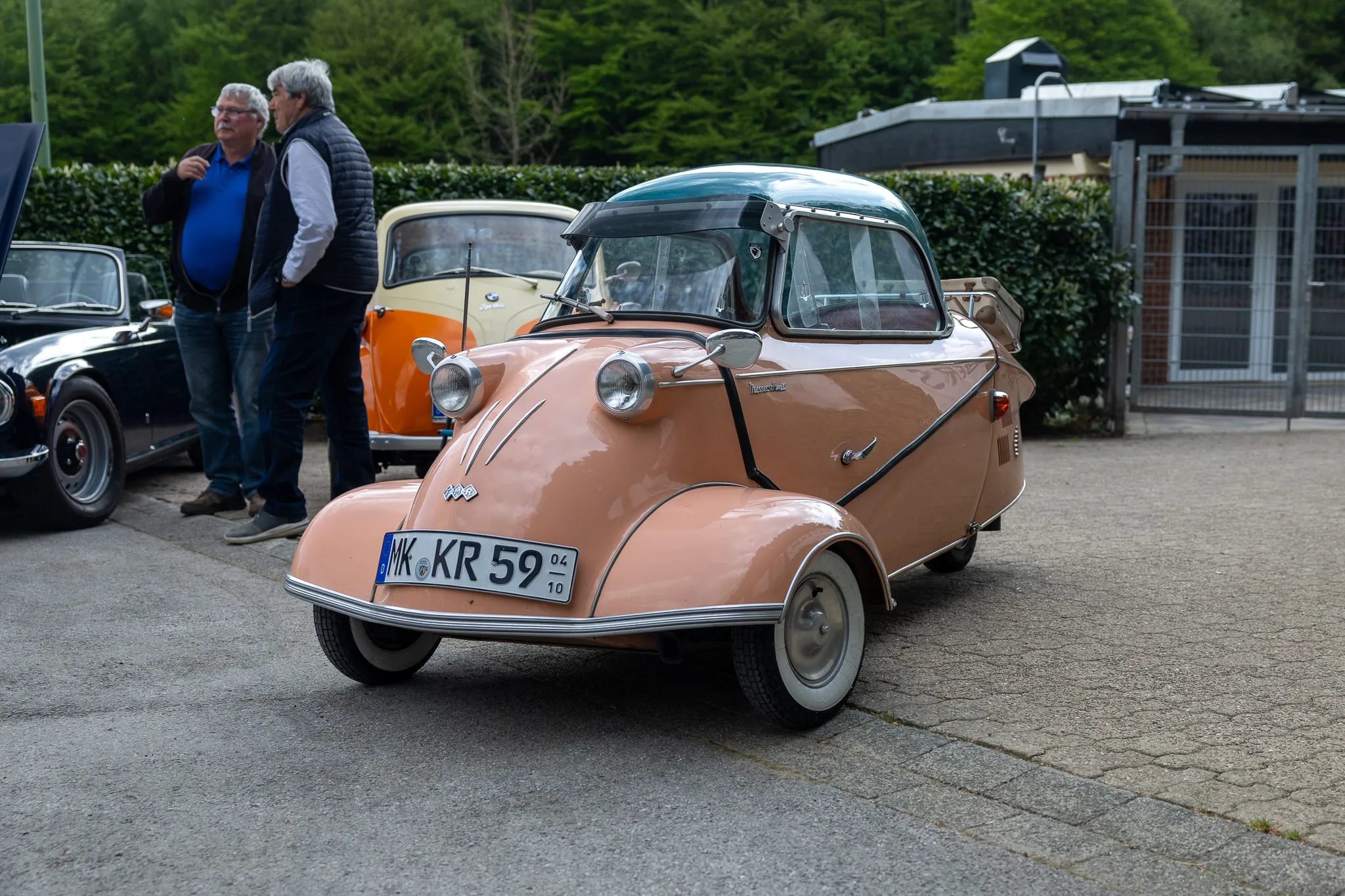 Rosafarbener BMW Isetta mit Glasdach steht auf einem Parkplatz, während ein Mann im Hintergrund steht und zusieht.