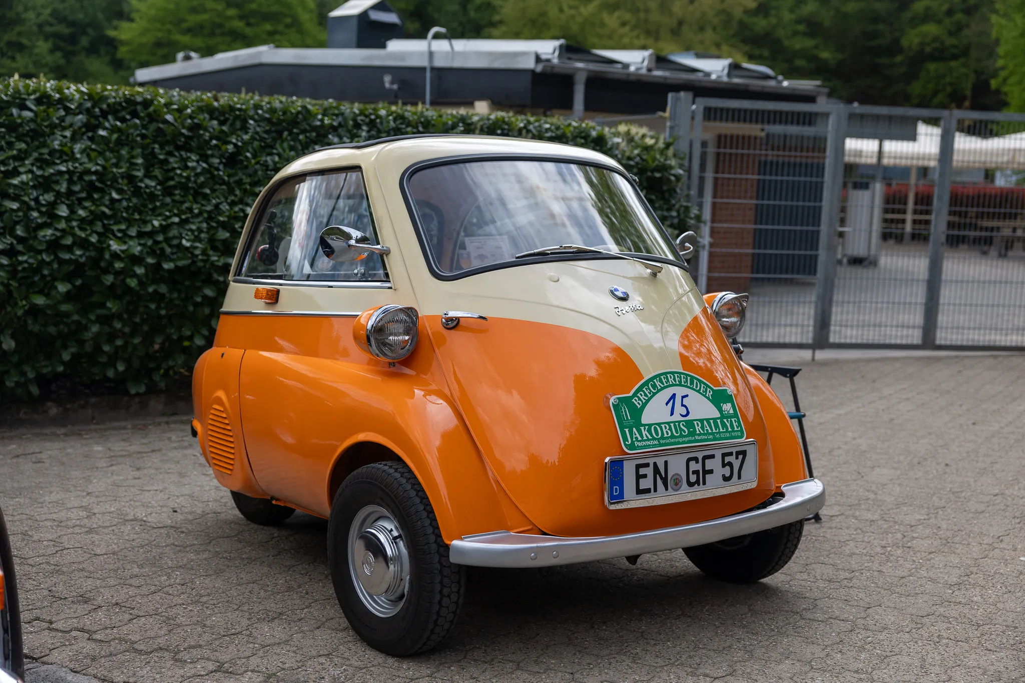 Ein orangefarbener BMW Isetta mit beigefarbenem Dach steht vor einer Hecke. Am Fahrzeug ist ein Aufkleber für die „Breckerfelder Jakobus-Rallye“ angebracht.
